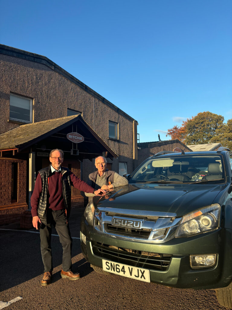 two men stand at the front of a pickup leaning on the bonnet. The Ritchie building is behind with the orange logo visible above them.